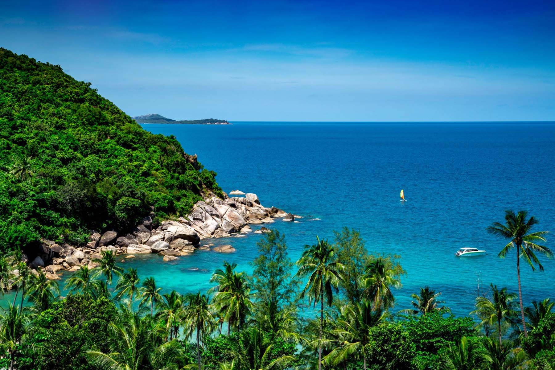 A turquoise cove on Koh Samui island, with palm trees, granite rocks, and a sailboat on the water